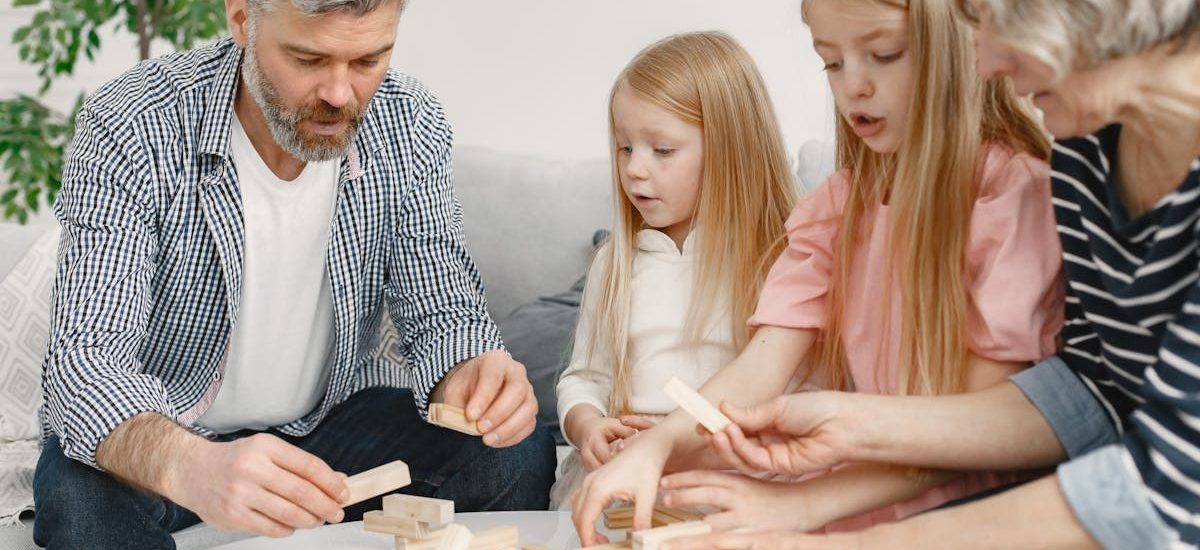 Four family members of different ages engage with toy blocks on a table indoors.